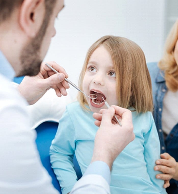 A dentist looking at a little girl’s smile while the mother sits nearby