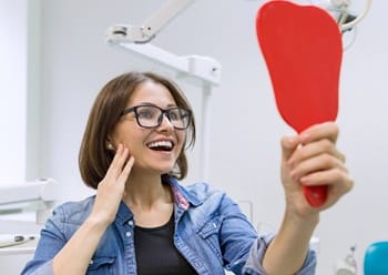 A middle-aged woman looking at her smile in the mirror after receiving custom-made dental crowns